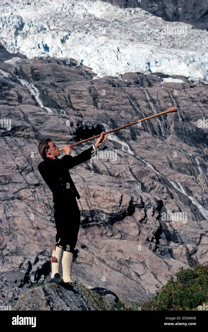 Ein junger Norweger Mann in traditioneller Kleidung vergangener Tage weht ein Vintage Holz Alphorn unter einem Berg Gletscher in Fjærland, Norwegen. Stockfoto