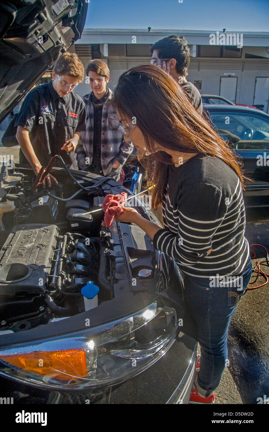 Schutzbrille tragen ein Teenager-Mädchen prüft ein Motorölstand, während ihre männlichen Klassenkameraden eine im Auto-laden-Klasse Batterie. Stockfoto