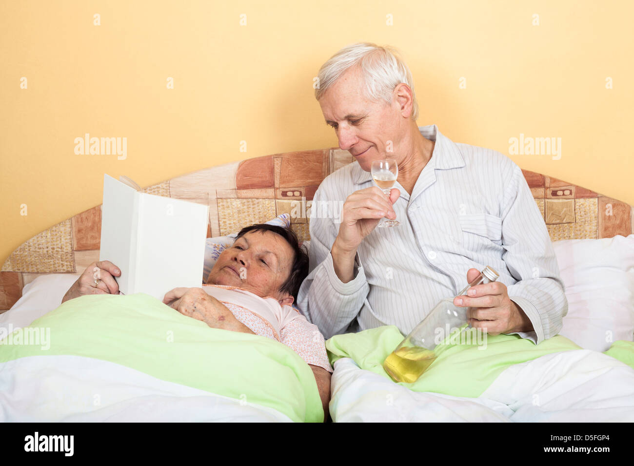 Lustiger senior Mann trinken Alkohol und Frau liest Buch im Bett Stockfoto