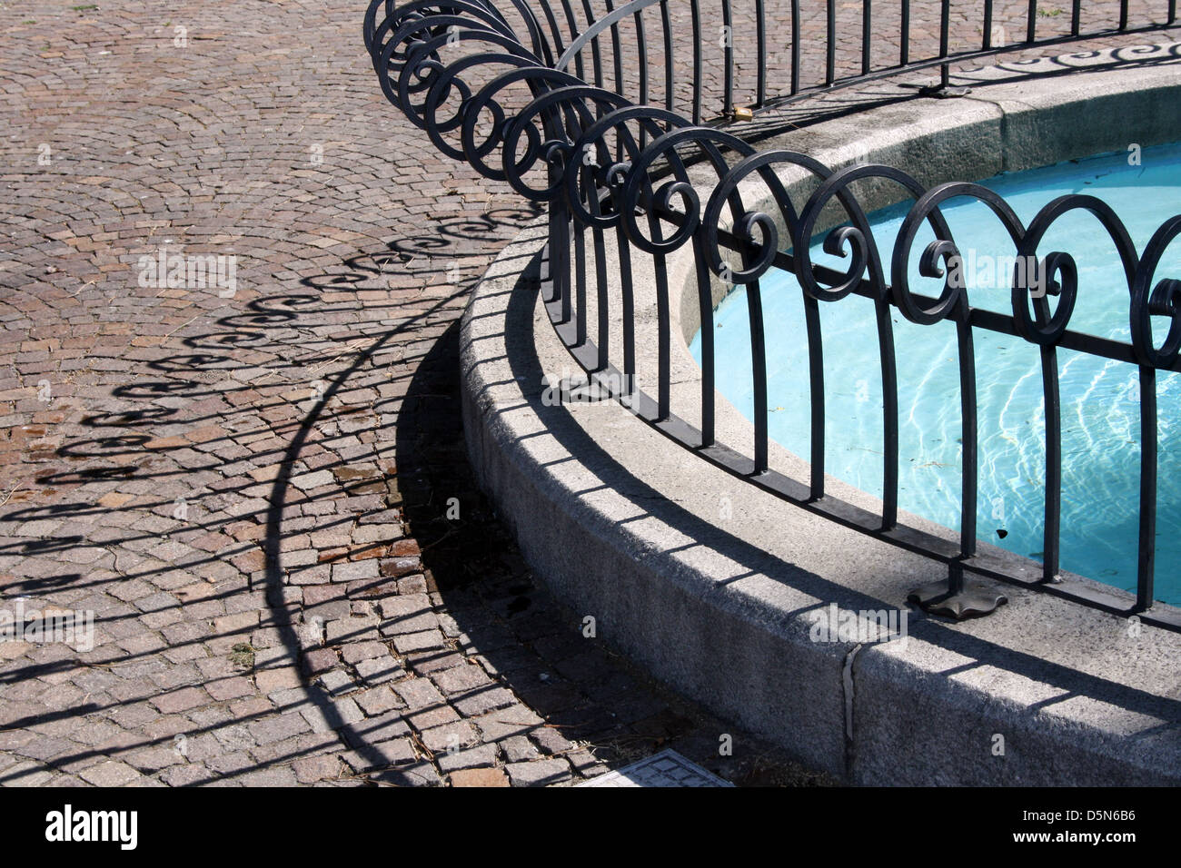Ein Halbkreis aus dekorativen gegossen Eisengitter um einen Brunnen, markante Schatten auf das Pflaster. Stockfoto
