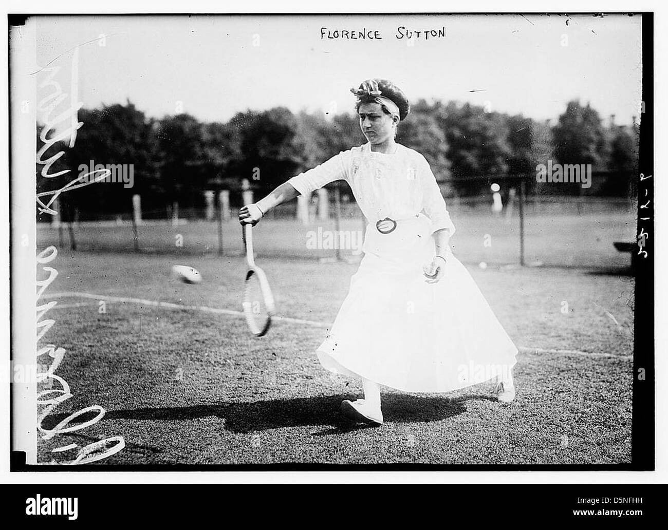 Ein Foto von Florence Sutton, einem US-amerikanischen Tennisspieler, aufgenommen während der Meisterschaft 1911. Das Bild fängt sie in Aktion ein, indem sie eine Rückhand auf einem Rasenplatz ausführt, ein klassisches Tennisoutfit und einen Hut trägt. Stockfoto