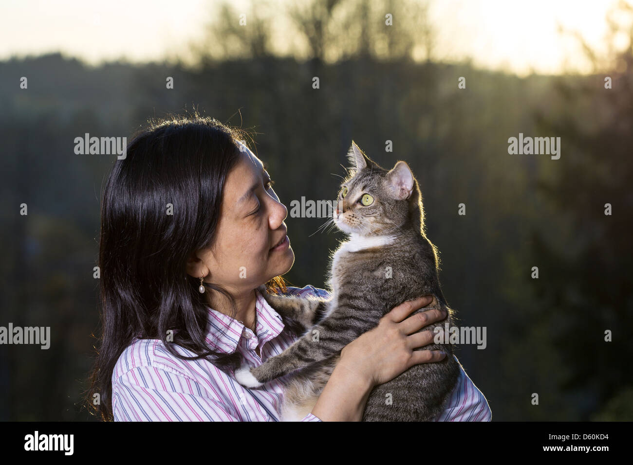 Horizontale Foto Reife Frau Holding Katze im Freien mit Fading Sonnenuntergang und Bäume im Hintergrund Stockfoto