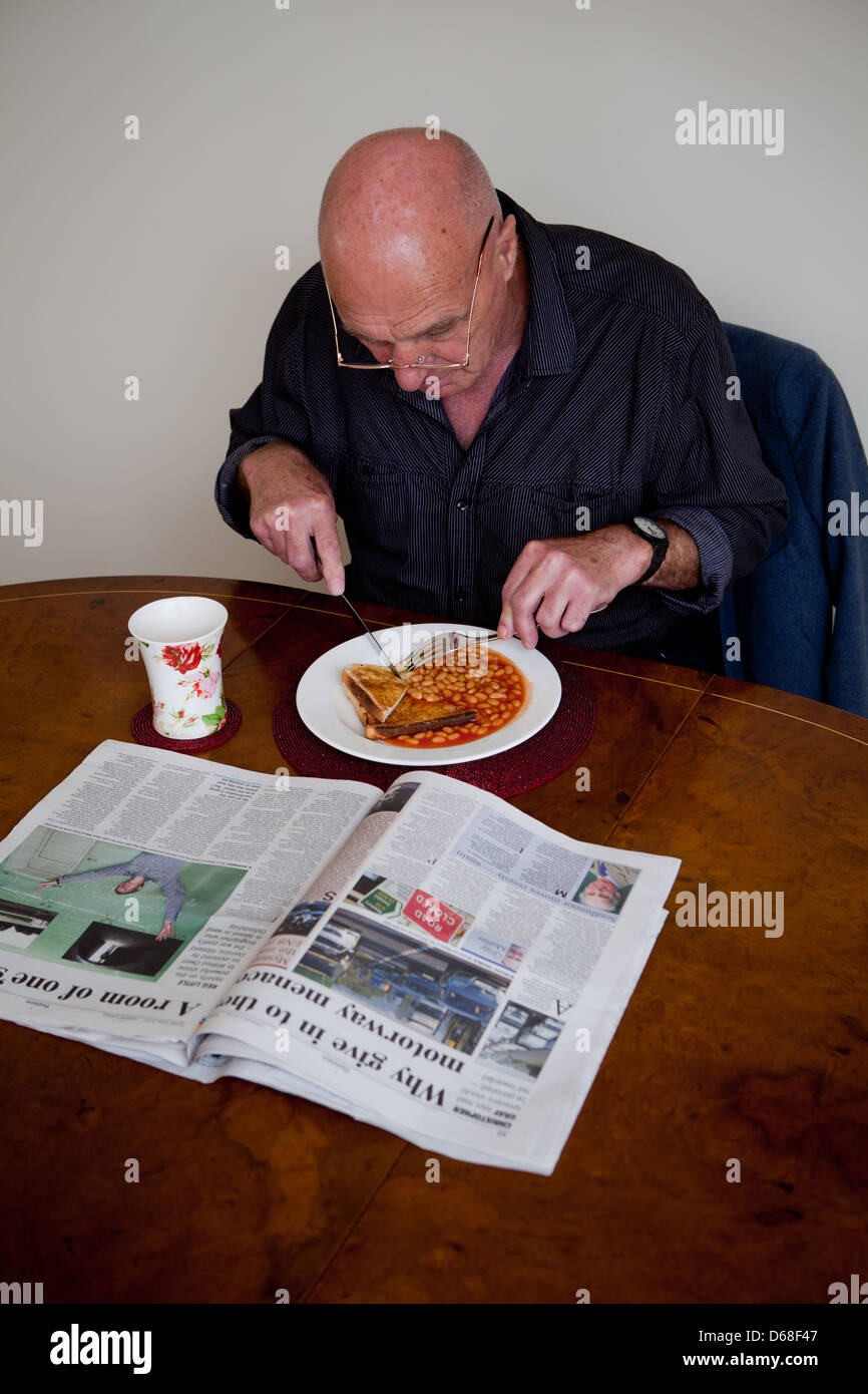 Ältere Mann an einem Tisch sitzen, Essen Bohnen auf Toast und liest eine Zeitung. Stockfoto