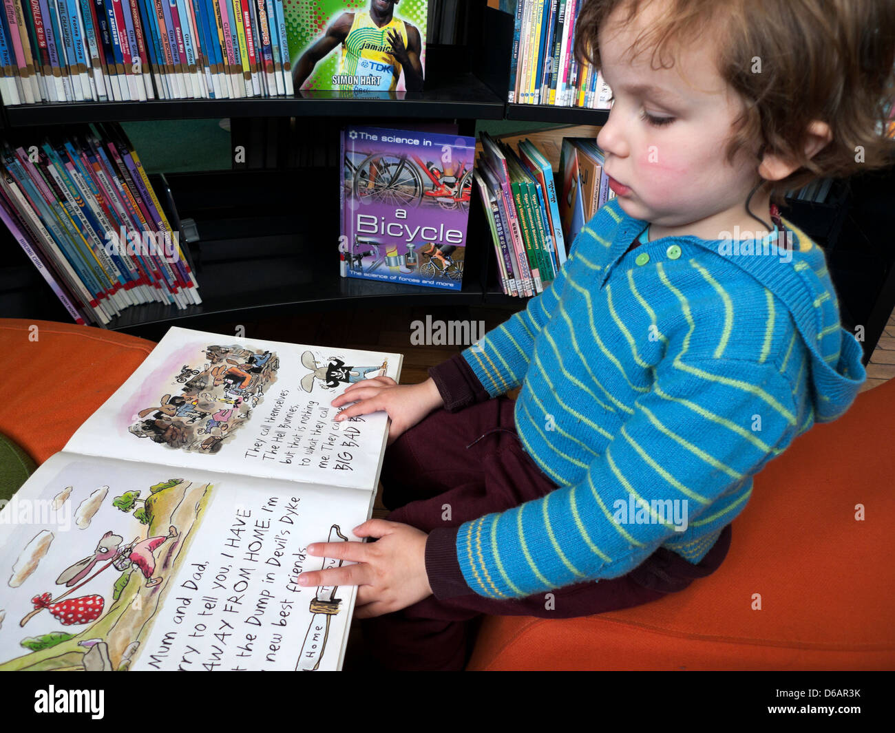 Ein 3 Jahre alter Junge Kind Lesen eines Tony Ross Bild Buch sitzend durch ein Regal in der Bibliothek Leseecke in Wales UK KATHY DEWITT Stockfoto
