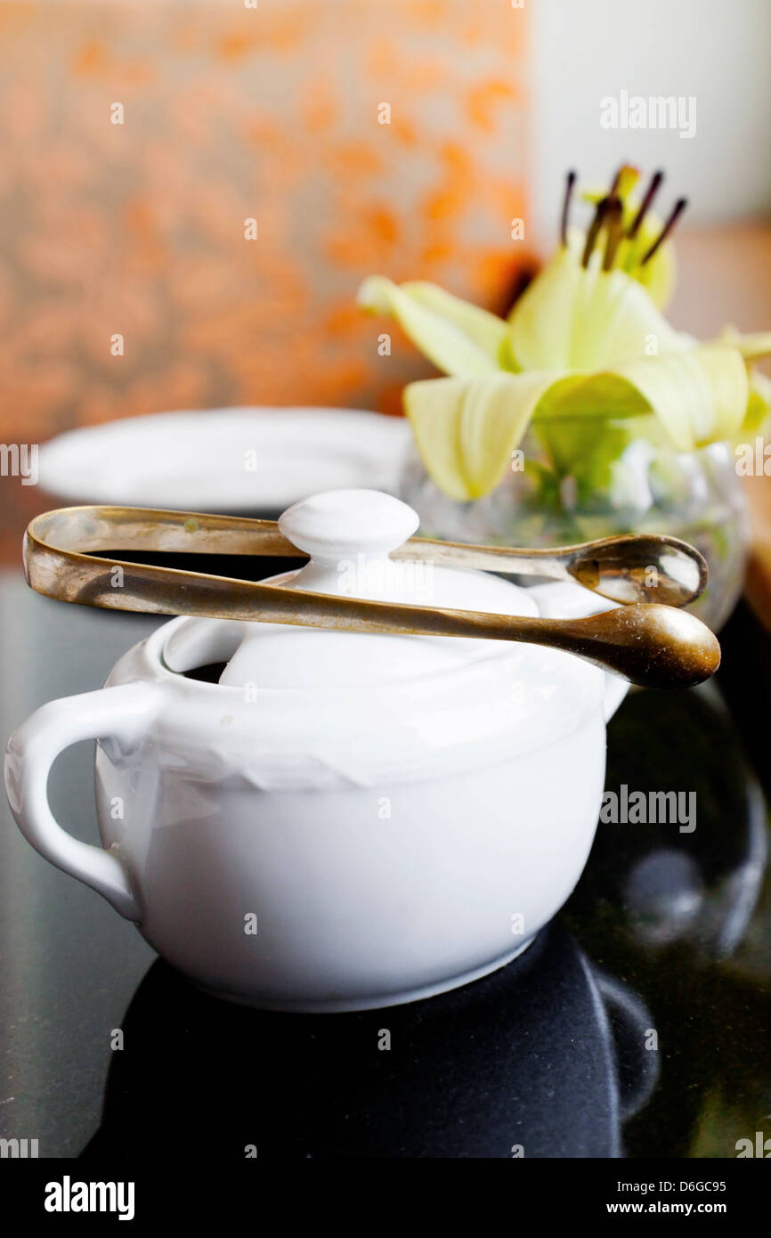 Vertikale Innenraum Detail des schwarzen Tisch mit Tee und Kaffee Geschirr Zuckerdosen und Knospe Vase mit einer Lilie durch ein Fenster Stockfoto