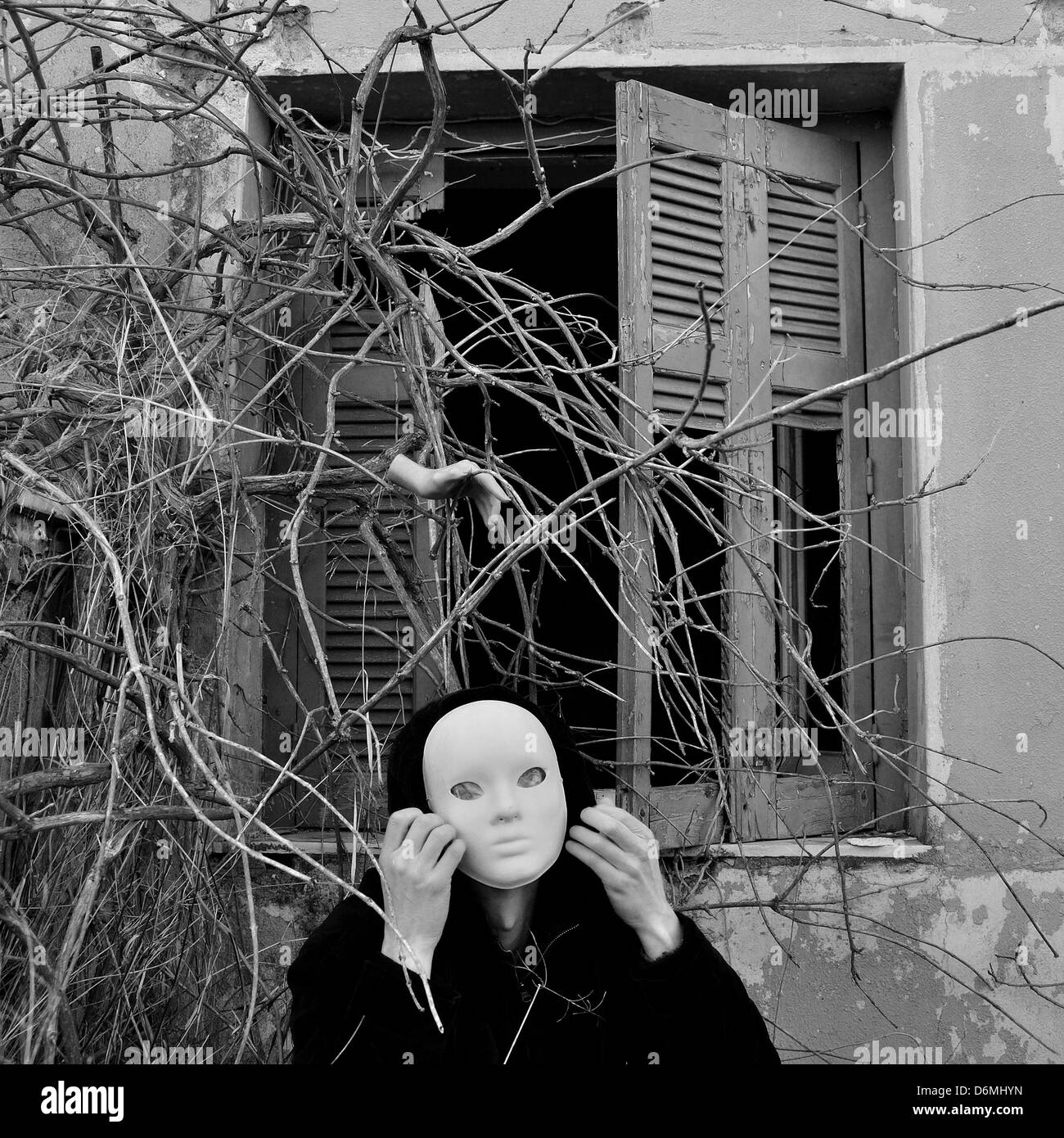 Figur mit weißer Maske unter Fenster mit verwilderten Pflanzen und Puppe Hand. Schwarz und weiß. Stockfoto