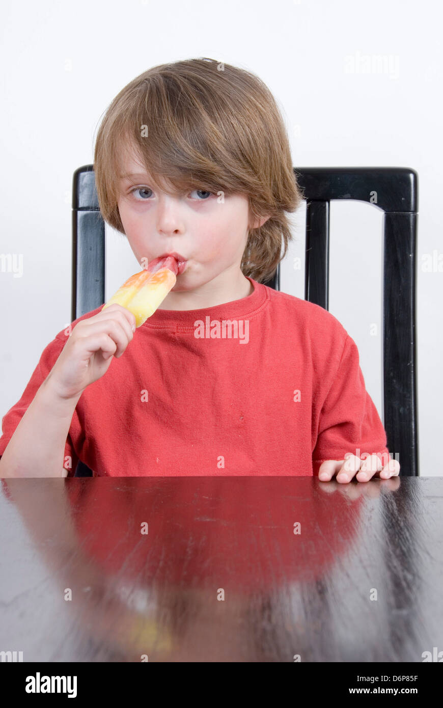 Kleiner Junge am Tisch saugen an einem bunten Eis am Stiel, Studioportrait isoliert auf weiss Stockfoto