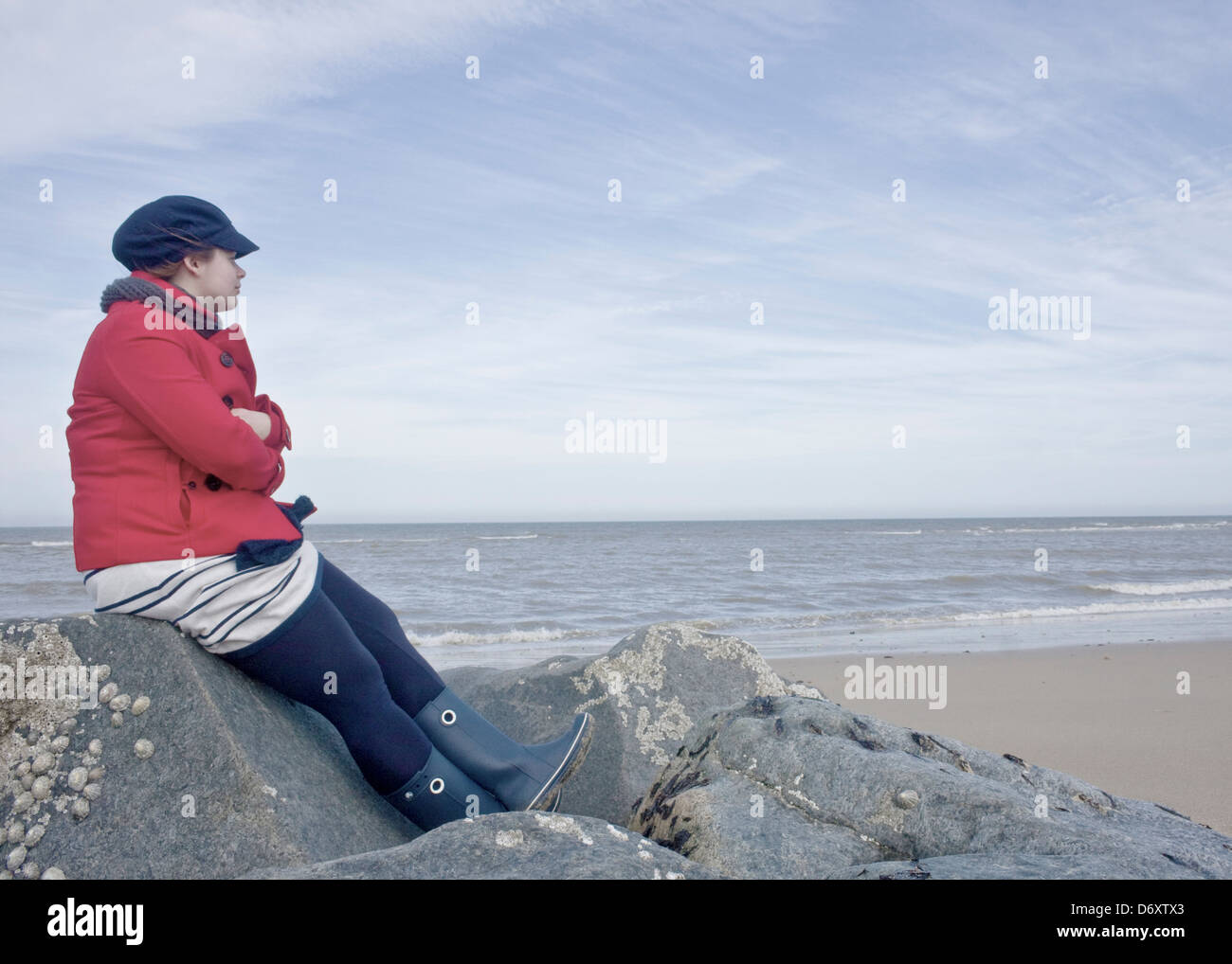 Frau im roten Mantel, sitzen auf den Felsen am Strand, Blick auf das Meer nachdenklich. Stockfoto