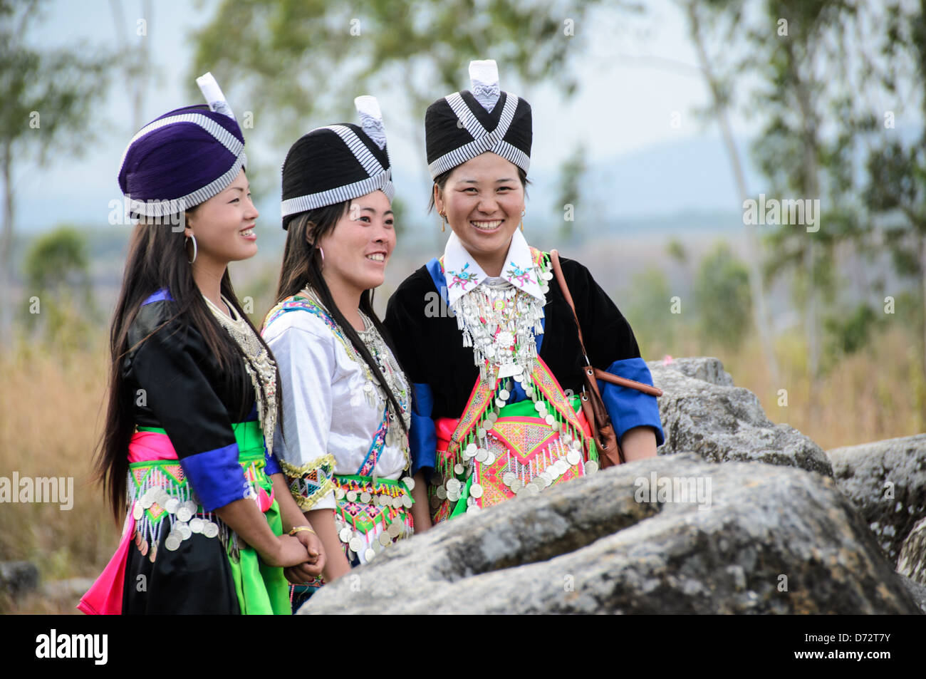 Hmong-Frauen in traditioneller Kleidung mit Silberschmuck Ebene der Krüge Laos | EBEINE DER KRÜGE, Laos — Drei Hmong-Frauen in traditioneller Neujahrskleidung versammeln sich neben einem megalithischen Steinbehälter an Stätte 1. Die schweren silbernen Münzketten, die sie tragen, dienen traditionell als mobile Familienspeicher, die während des jährlichen Festivals auffällig zur Schau gestellt werden. Das geschnitzte Steingefäß hinter ihnen gehört zur weitläufigen Bestattungslandschaft von Thong Hai Hin, wo mehr als 300 Krüge auf dem offenen Plateau stehen — Teil eines UNESCO-Weltkulturerbestandes, der 2019 eingetragen wurde und durch kürzliche Ausgrabungen nun als f Stockfoto