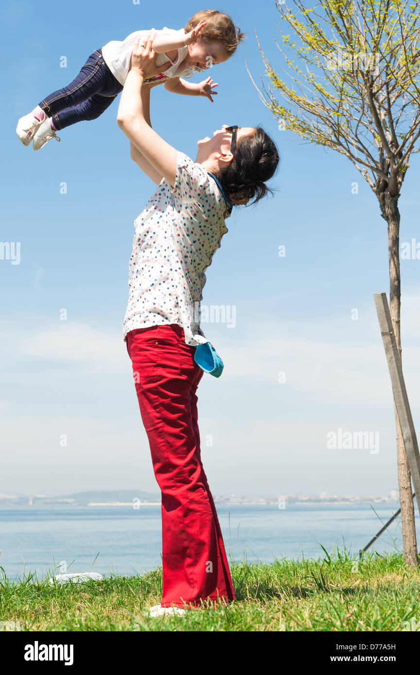 Glückliche Mutter hob ihr kleines Mädchen zu Frühling Gras Himmel Stockfoto