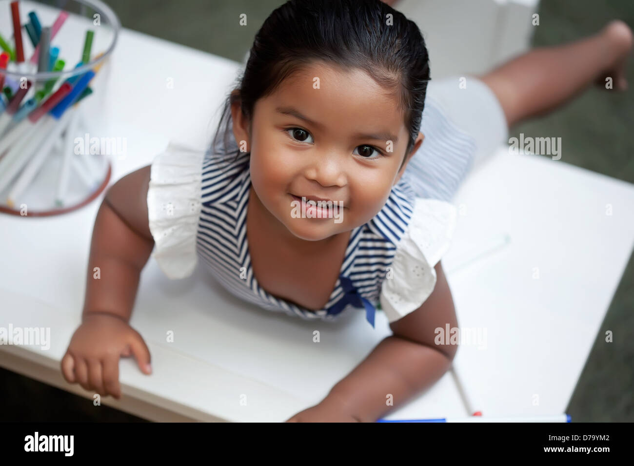 Latino Mädchen mit einem Lächeln legt sich auf weiße Vorgangstabelle mit Markern einfärben Stockfoto