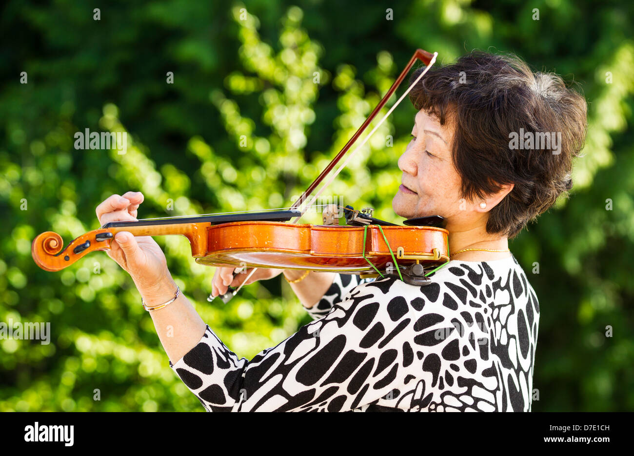 Horizontale Foto von Senior Asiatin vor seitlich bei der Geige im Freien mit grünen Bäumen im Hintergrund Stockfoto