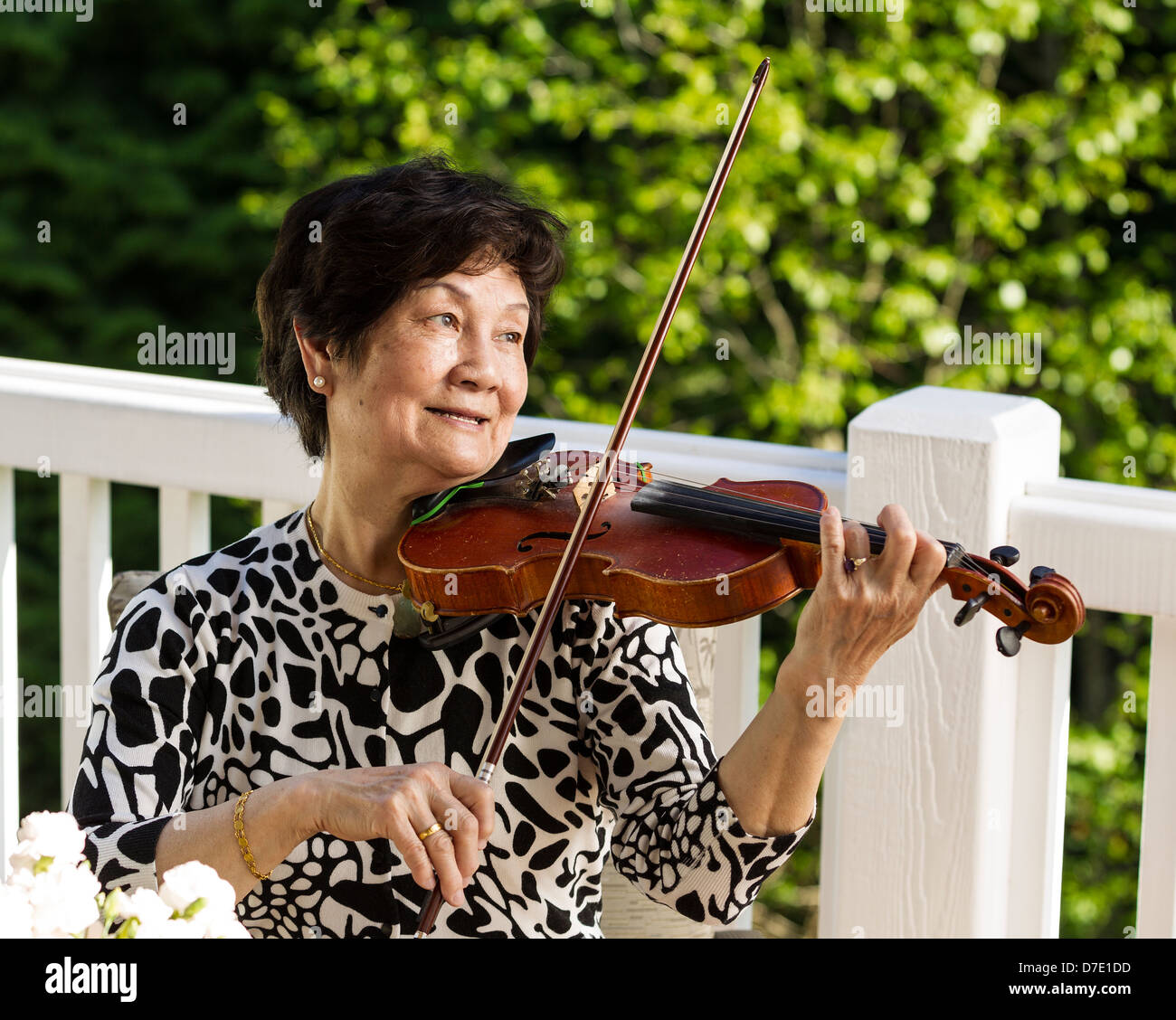 Horizontale Foto Senior asiatische Frau sitzen beim Spielen der Violine im Freien mit grünen Bäumen im Hintergrund Stockfoto