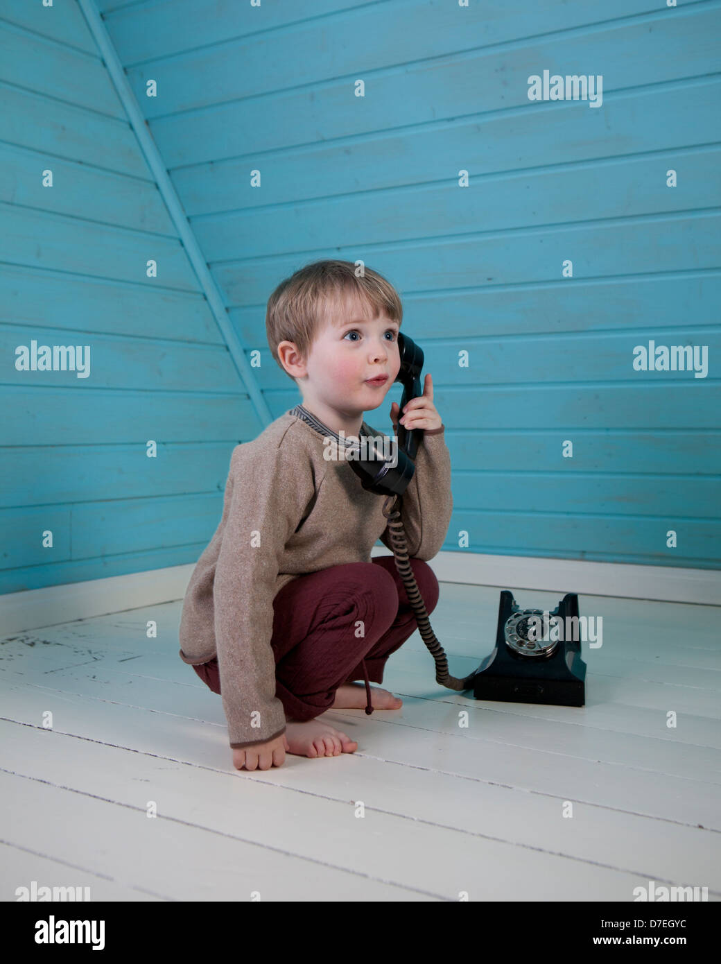 Ein Junge spielt mit einem alten altmodischen Wählscheibe Telefon sitzen auf dem weißen Holzboden barfuß in einem blauen Loft-Zimmer Stockfoto