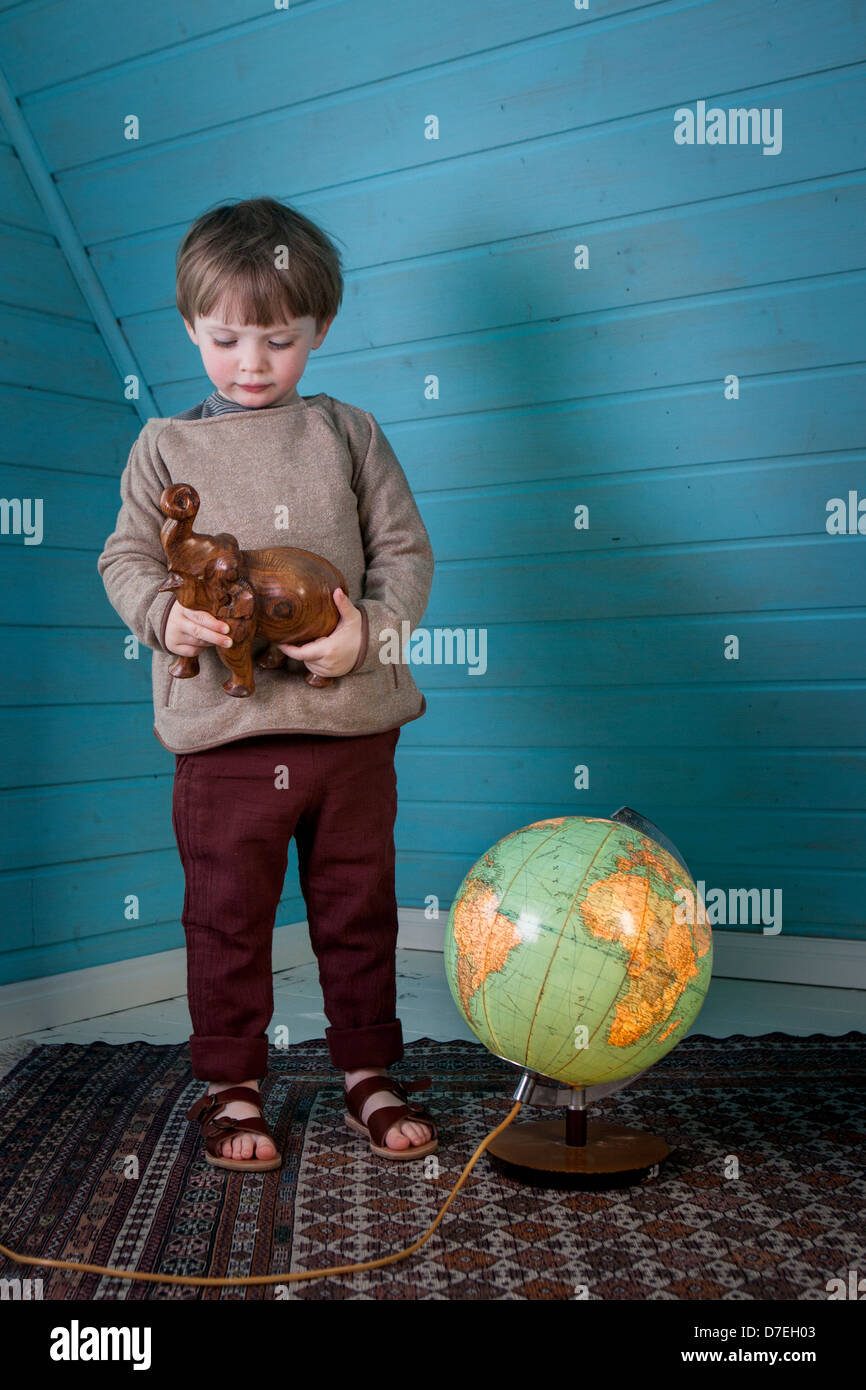 Kleiner Junge betrachtet man eine aus Holz geschnitzten Elefanten in seinen Händen stehen neben einem beleuchteten Globus zeigt Afrika und Südamerika Stockfoto