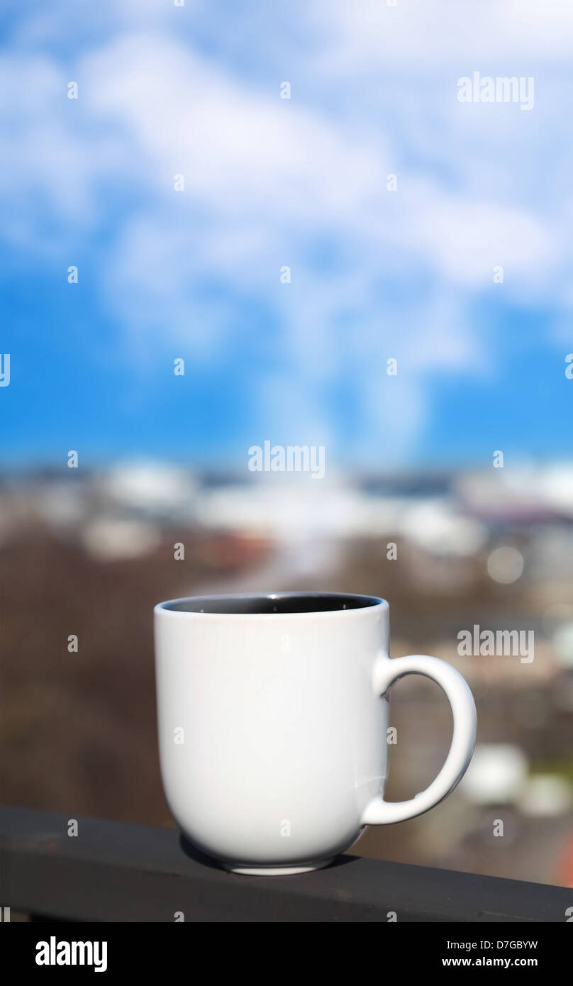 Weiße Tasse steht auf Balkongeländer über verschwommenes Stadtbild mit Dampf sieht aus wie Wolken am blauen Himmel Stockfoto