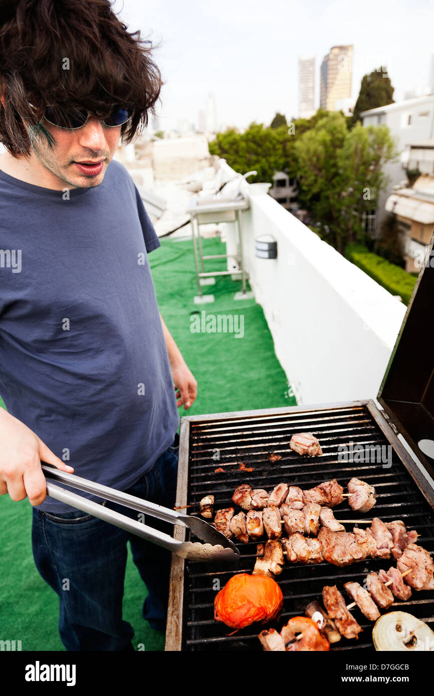 Frühen 30er Jahren kaukasischen Mann beschäftigt Grillen etwas Fleisch und Gemüse auf dem Dach eines Gebäudes in einer städtischen Umgebung. Stockfoto