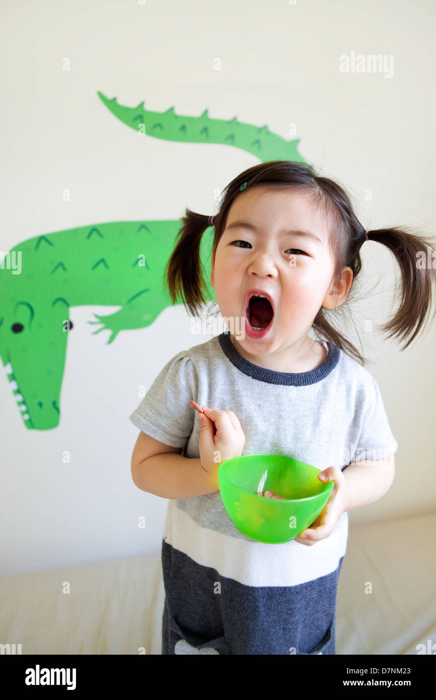 Korean American Kleinkind in Zöpfen zu essen Snacks aus der grünen Schale und Krokodil Wandkunst hinter Kamera schreien. Stockfoto