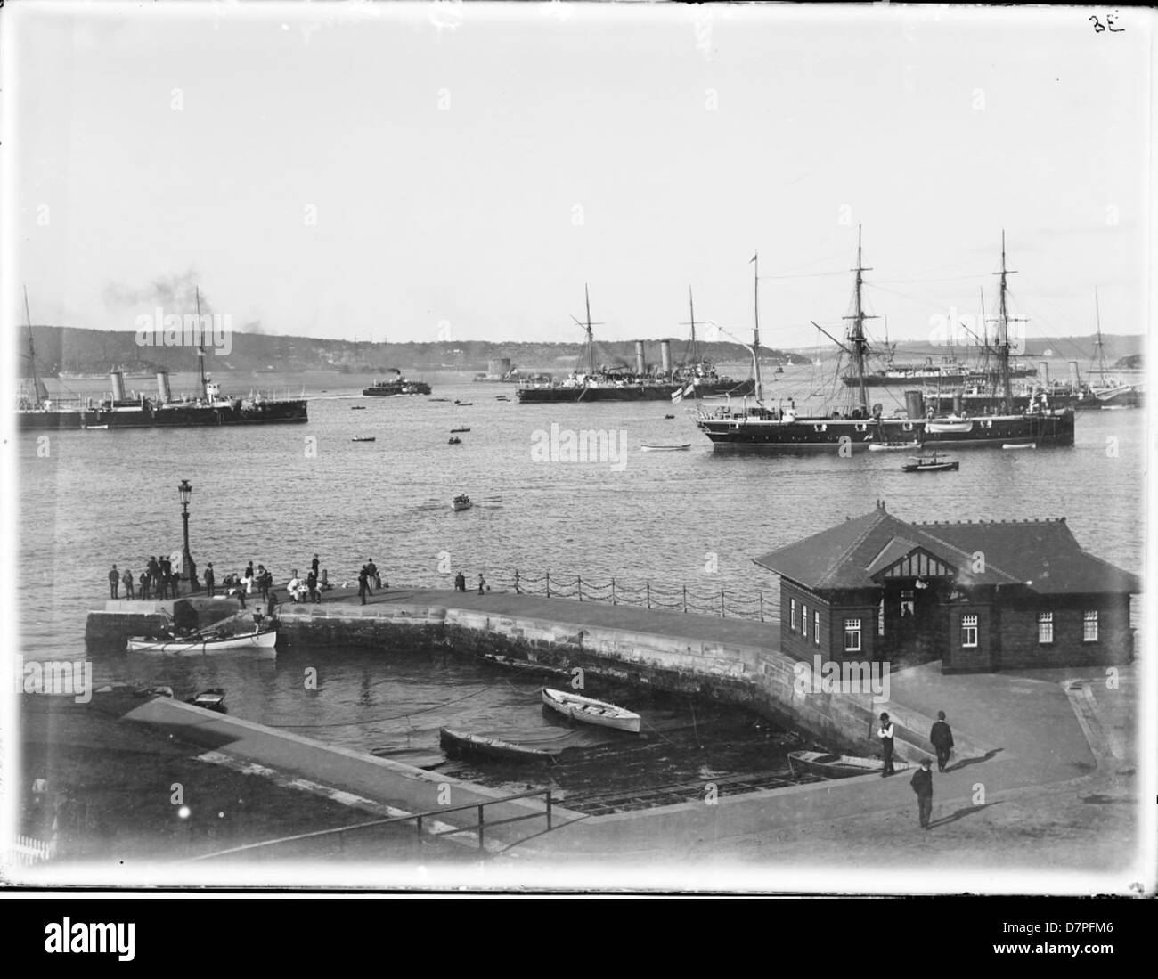 Ein Schwarzweiß-Foto, das eine Fähranlegestelle im Hafen von Sydney zeigt, mit der HMS Royalist, ein Mann o' war, in der Nähe angedockt. Das Bild zeigt die historische Uferpromenade von Sydney, einschließlich Fähren, Booten und einer Reihe von Häusern entlang des Hafens. Die Anlegestelle ist ein wichtiger maritimer Ort in Sydneys Geschichte. Stockfoto