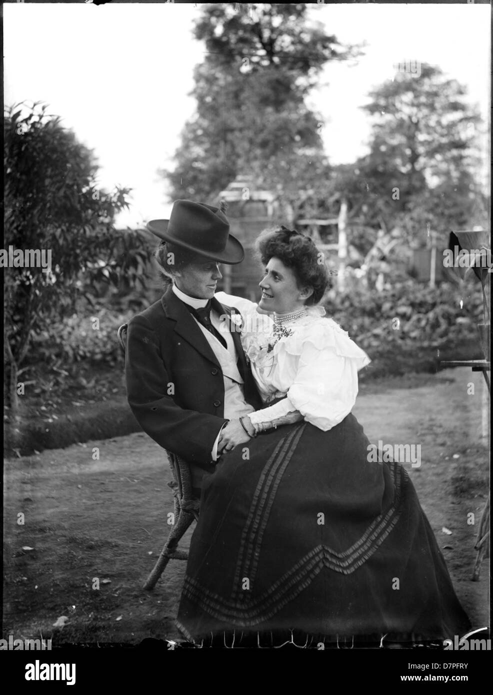 Ein junges Paar, das in einem Garten sitzt, in historischer Kleidung, die die Modetrends des späten 19. Jahrhunderts widerspiegelt, auf einem retuschierten Foto aus der Zeit von 1885. Stockfoto