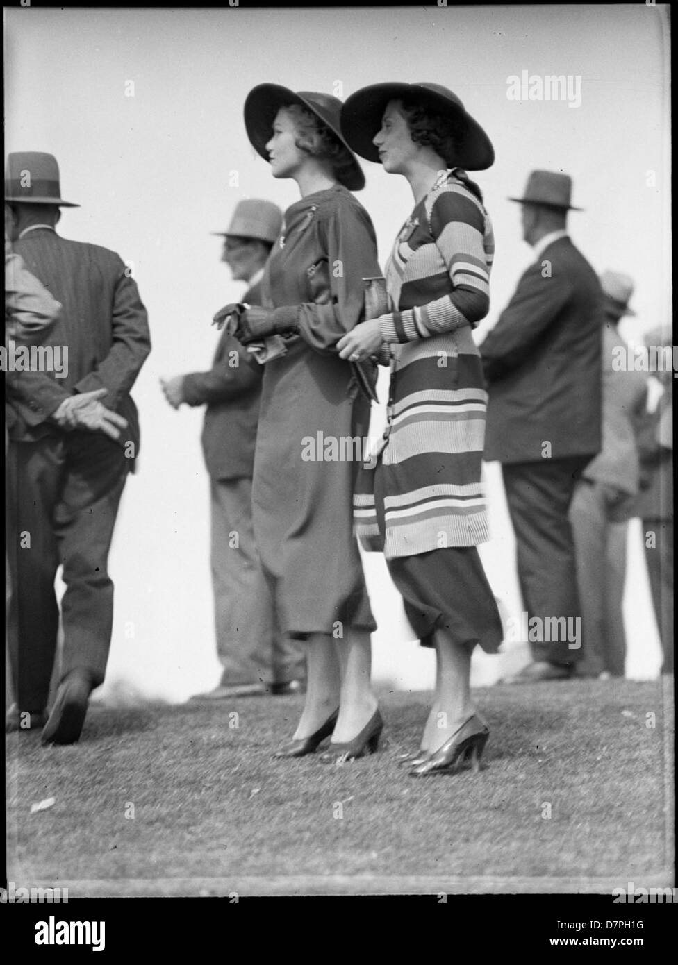 Dieses Foto aus den 1930er Jahren zeigt Rennfahrer auf dem Warwick Farm Racecourse in Australien und zeigt die Modetrends dieser Zeit. Das Bild spiegelt die stilvolle Kleidung wider, die sowohl Männer als auch Frauen beim Pferderennen getragen haben. Stockfoto