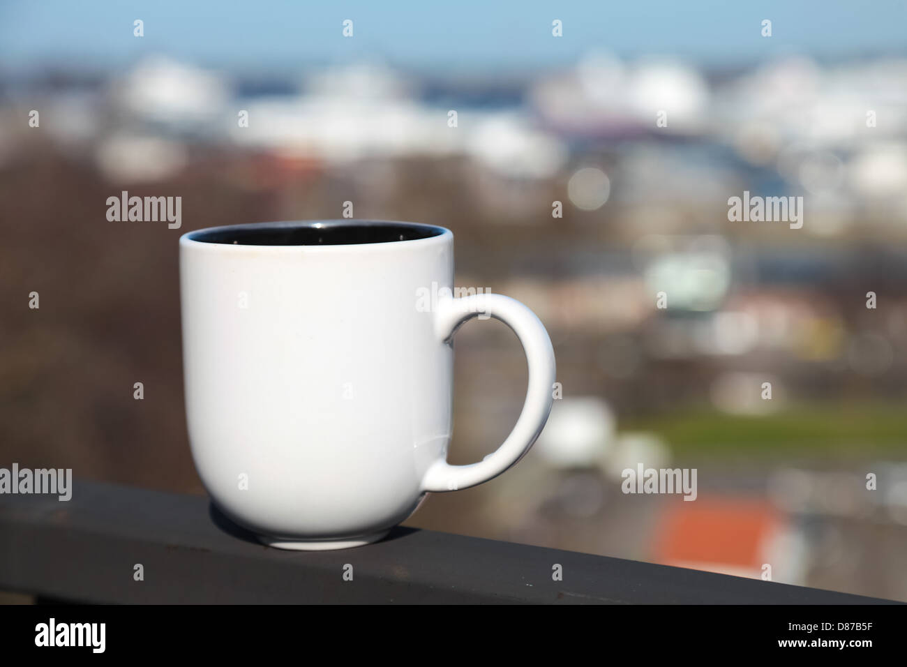 Weiße Tasse Kaffee steht am Balkongeländer über verschwommenes Stadtbild Stockfoto