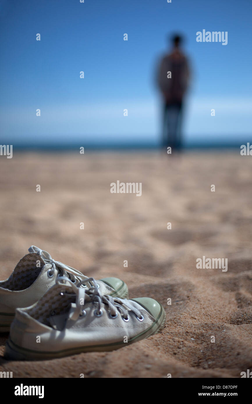 Ein Paar Turnschuhe auf den Strand und eine Person zu Fuß in einem Abstand zum Wasser an einem sonnigen Tag am Strand. Stockfoto