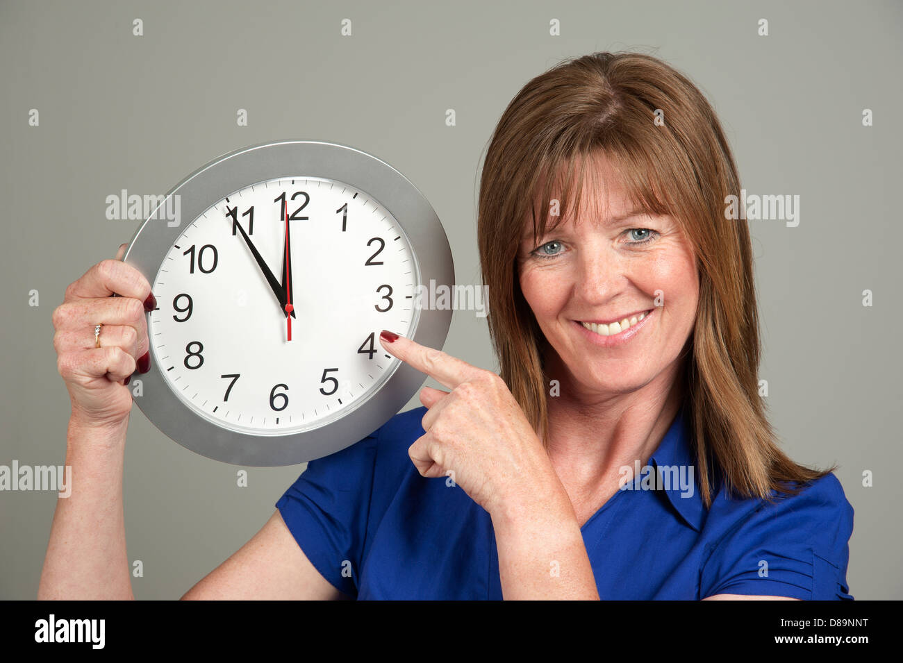 Frau mit überrascht Ausdruck hält eine Uhr mit der Zeit auf fünf vor zwölf Stockfoto