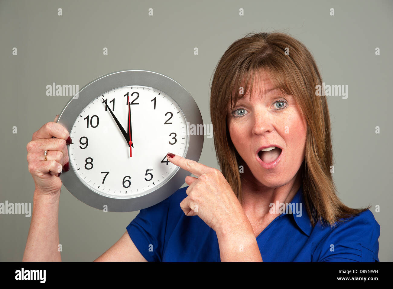 Frau mit überrascht Ausdruck hält eine Uhr mit der Zeit auf fünf vor zwölf Stockfoto