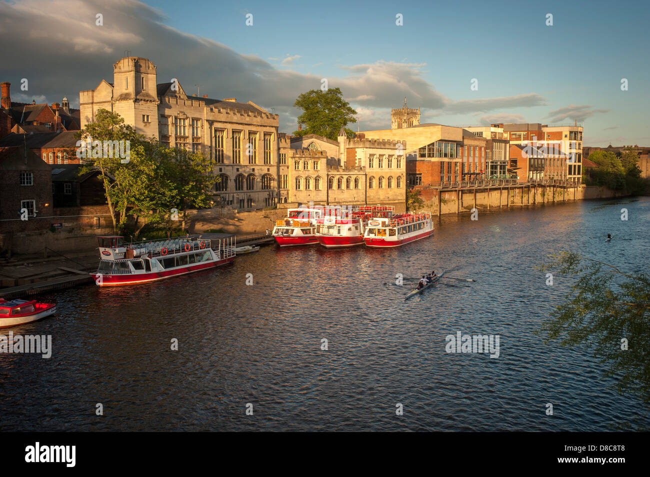 Eine Flotte von Kreuzfahrt- und Touristenbooten, die an einem sonnigen Sommerabend an der Guildhall mit einem Ruderboot im Vordergrund vor Anker liegt. River Ouse, York. Stockfoto