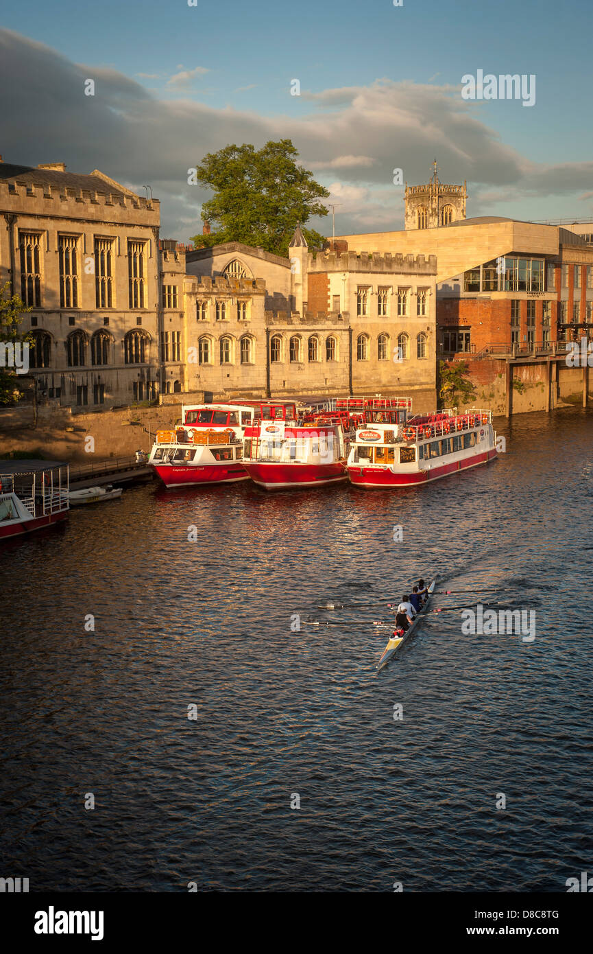 Eine Flotte von Kreuzfahrt- und Touristenbooten, die an einem sonnigen Sommerabend an der Guildhall mit einem Ruderboot im Vordergrund vor Anker liegt. River Ouse, York. Stockfoto