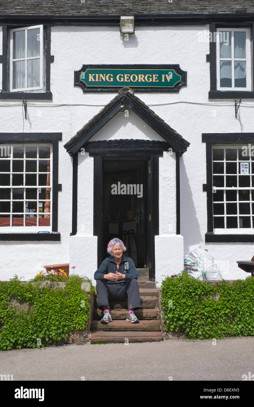 Ältere Frau sitzt vor dem König George IV Pub, Eskdale Green, West Cumbria, England UK Stockfoto
