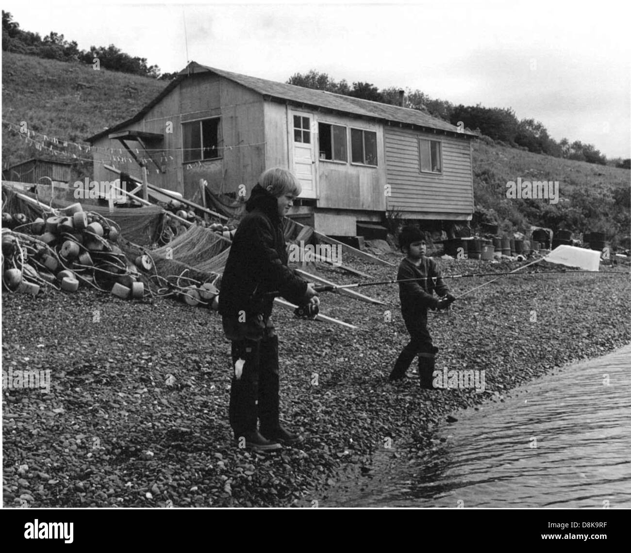 Zwei Jungen fischen in einem See oder Fluss in der Nähe einer alten Holzhütte, umgeben von einer rustikalen und natürlichen Umgebung im Freien. Stockfoto