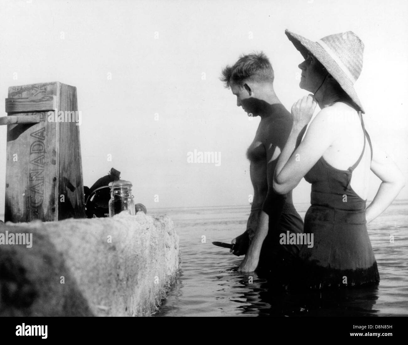 Dieses Vintage-Schwarzweiß-Foto zeigt einen Mann und eine Frau, die im Wasser stehen. Das Foto fängt einen Moment in der Zeit ein und spiegelt eine ruhige oder romantische Szene aus der Vergangenheit wider. Das Paar taucht in das Wasser ein, möglicherweise in einen Fluss oder See, und genießt einen friedlichen Moment zusammen. Stockfoto