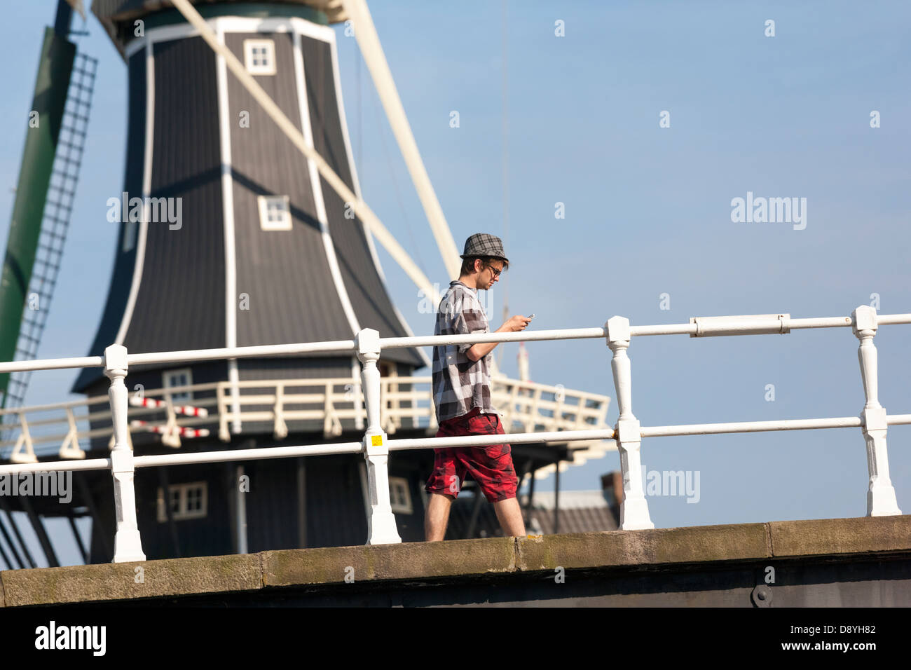 Junger Mann Überprüfung sein Smartphone zu Fuß auf einer Brücke vor Wahrzeichen Windmühle De Adriaan in Haarlem, Holland. Sommer. Stockfoto