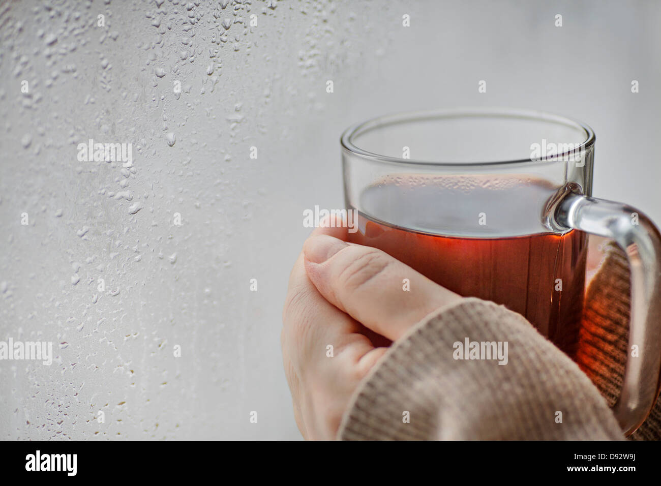 Menschliche Hände halten eine Tasse Tee einen Fensterplatz mit Kondenswasser drauf Stockfoto