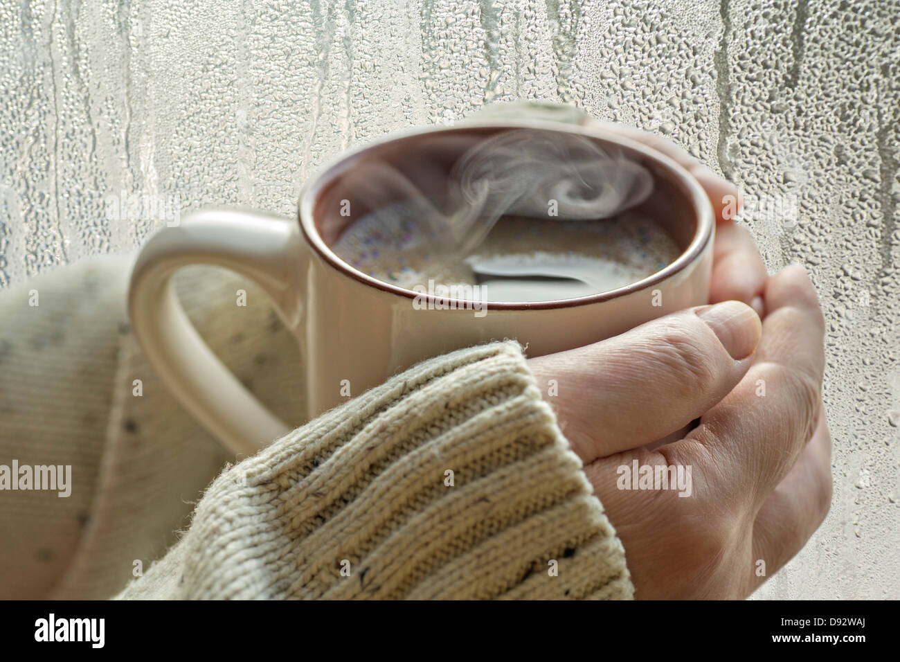 Menschliche Hände halten einen Becher mit Dampf aufsteigt, neben einem Fenster mit Kondensation darauf Stockfoto