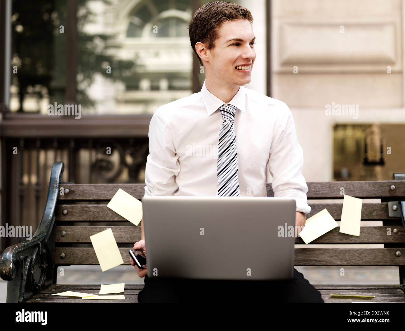 Lächelnde Geschäftsmann hält Smartphone während mit einem Laptop sitzen auf Bank mit Haftnotizen Stockfoto