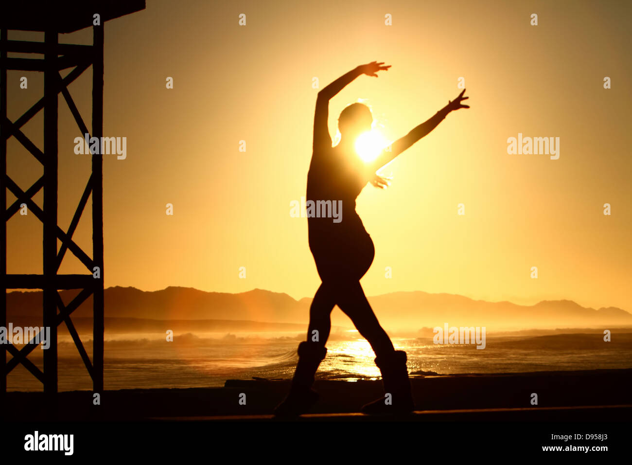 Eine schöne exotische Tänzerin ist durch die Sonne über dem Ozean Silhouette. Sie steht auf dem Pier in Port Alfred. Stockfoto