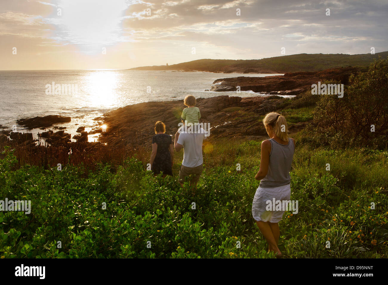 Einschließlich der Familiengruppe Vater tragen 3 jährige Shoudlers Spaziergänge nach unten in Richtung der felsigen Bucht bei Sonnenuntergang Stockfoto