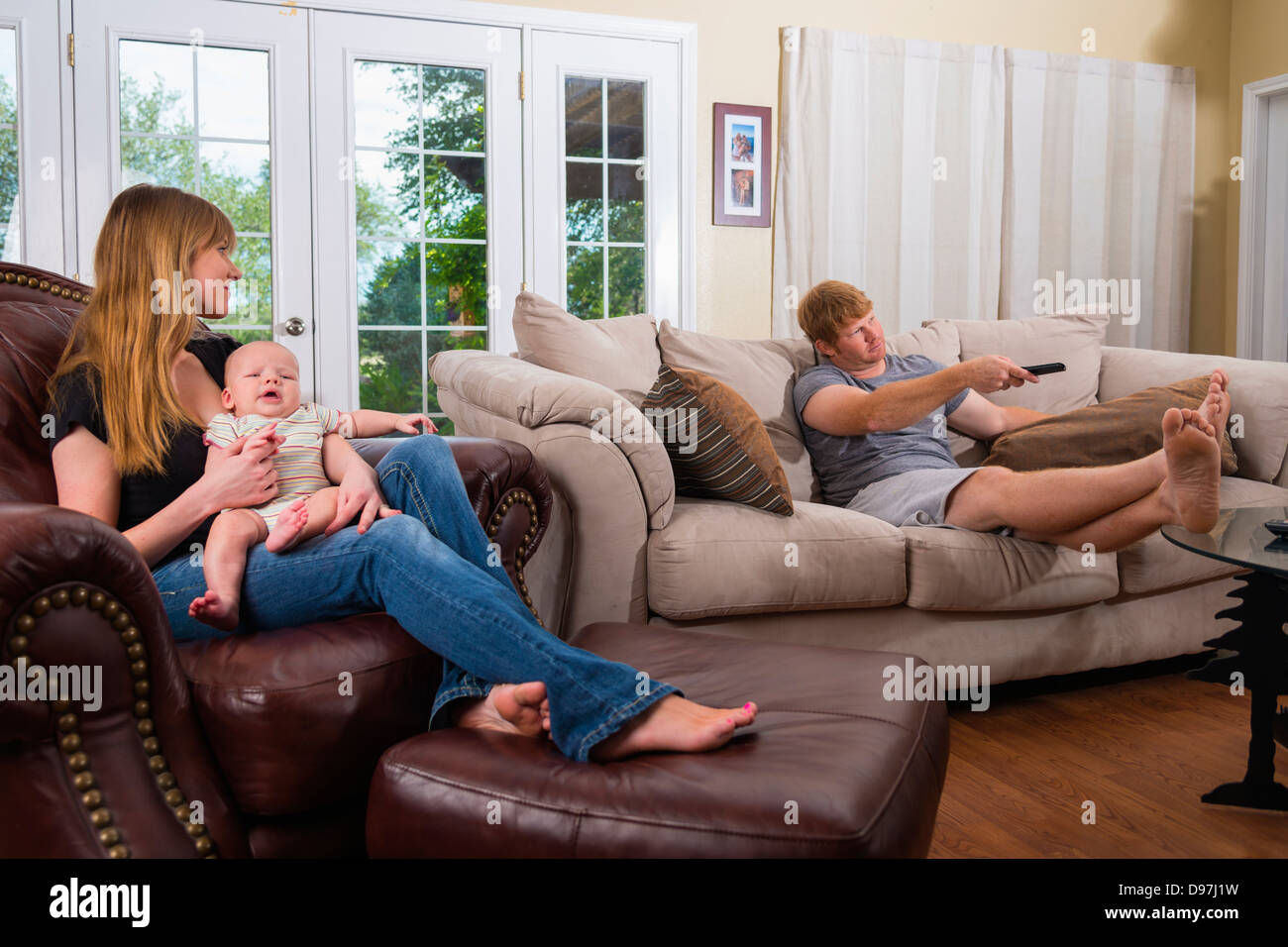 Junge Eltern - Mutter kümmert sich um Baby, während Vater in ihrem Haus Wohnzimmer vor dem Fernseher ist Stockfoto