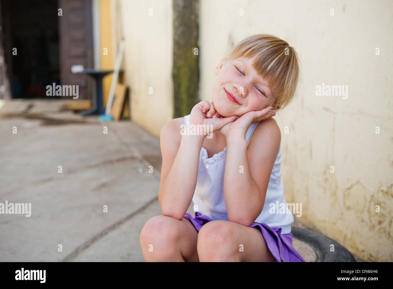 Porträt von süß lächelnde junge Mädchen mit Augen geschlossen vor Betonwand draußen sitzen Stockfoto