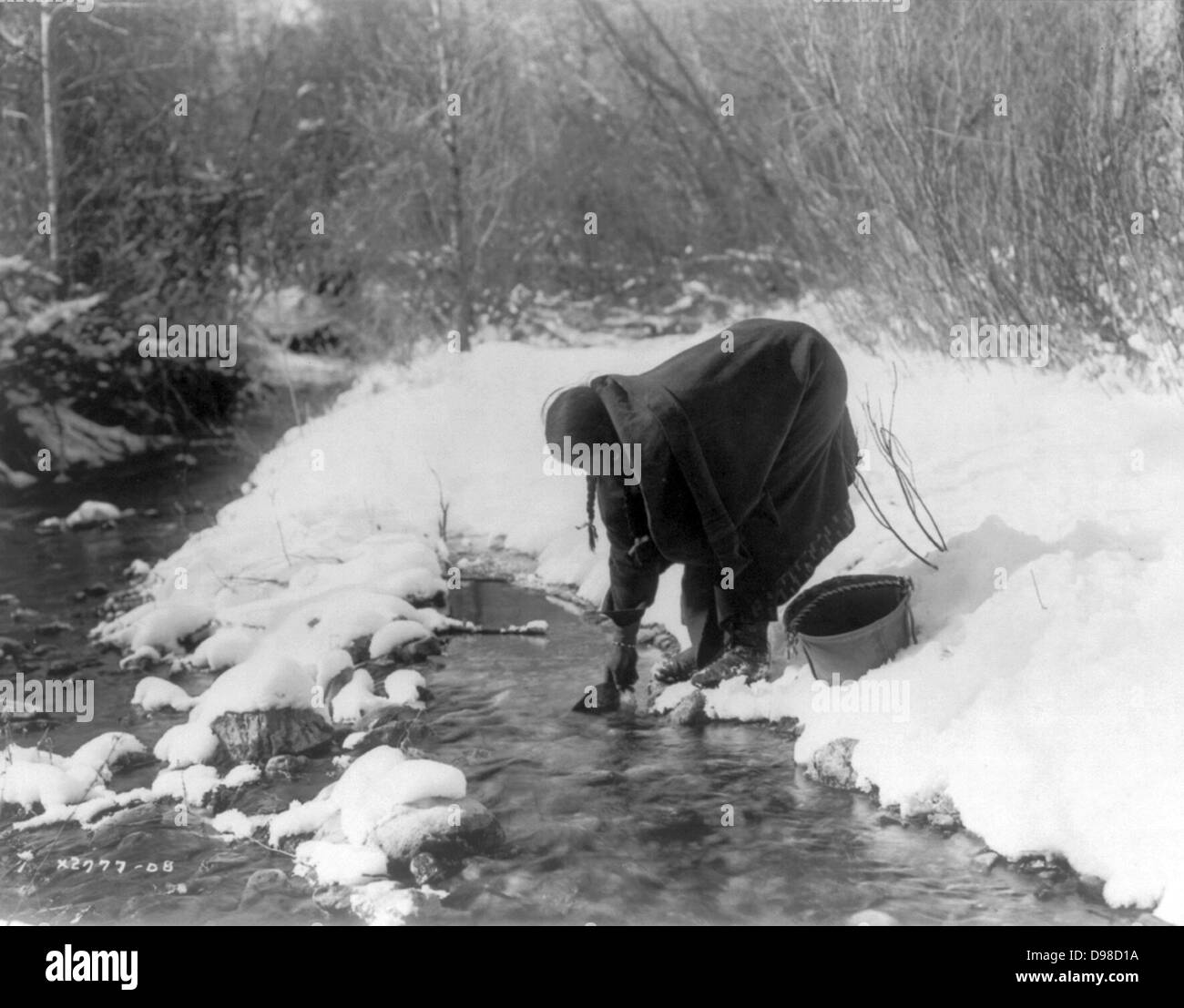 Apsaroke Frau im Schnee schöpfen Wasser aus einem Bach mit einer Dose Eimer neben ihr. Stockfoto