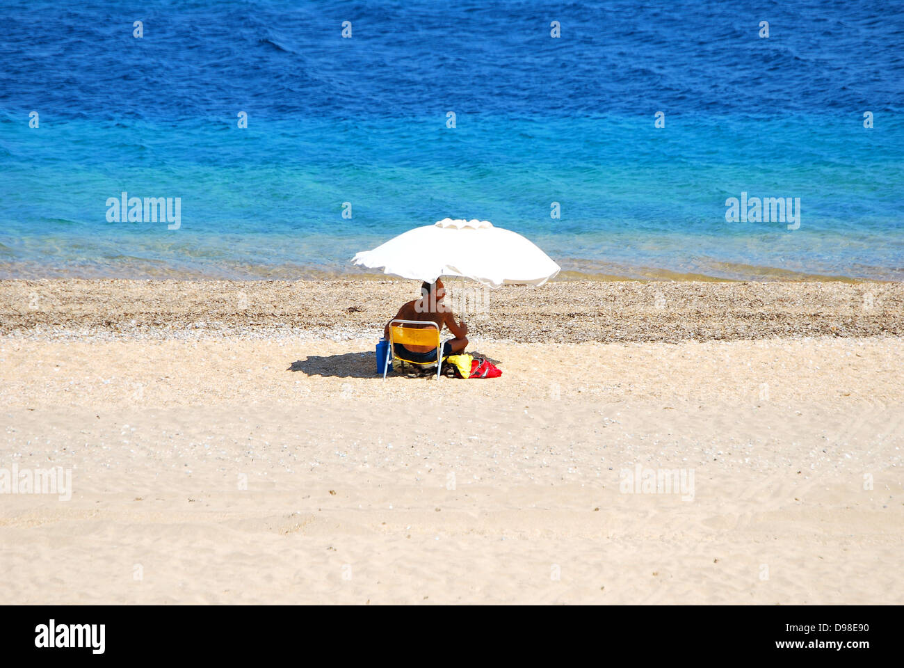 Menschenleeren Sandstrand mit einem einsamen männlichen Sonnenanbeter bergende unter einem Sonnenschirm mit türkisblauen Meer im Hintergrund. Stockfoto