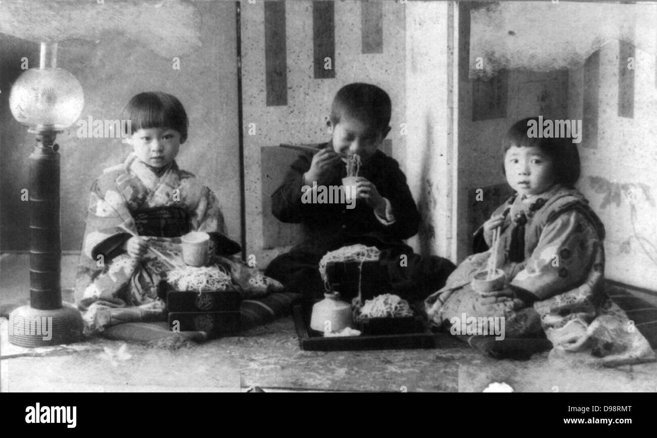 Drei japanische Kinder sitzen auf Kissen auf dem Boden, Nudeln mit Stäbchen zu essen. Anfang des 20. Jahrhunderts. Stockfoto