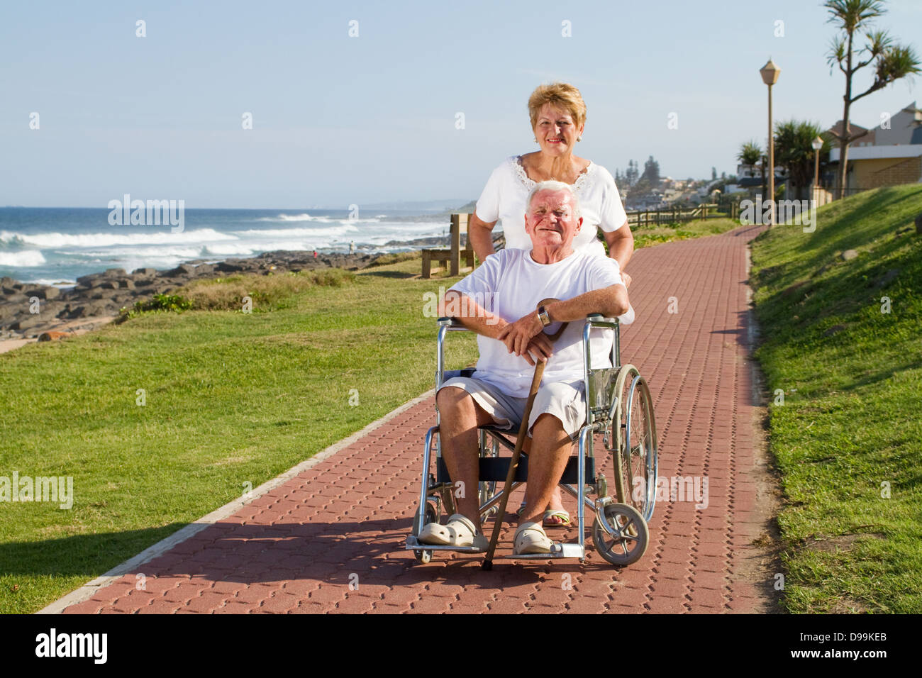 Behinderte senior Mann gedrängt an sonnigen Tag am Rollstuhl durch seine liebevolle Frau am Strand Stockfoto