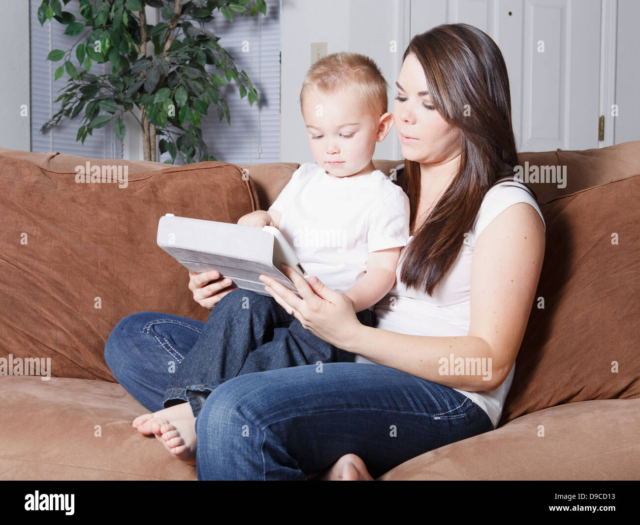 Hübsche junge kaukasischen Mutter und Sohn blondes Kleinkind sitzt zu Hause auf Sofa zusammen auf einem drahtlosen mobilen Tablet lesen. Stockfoto