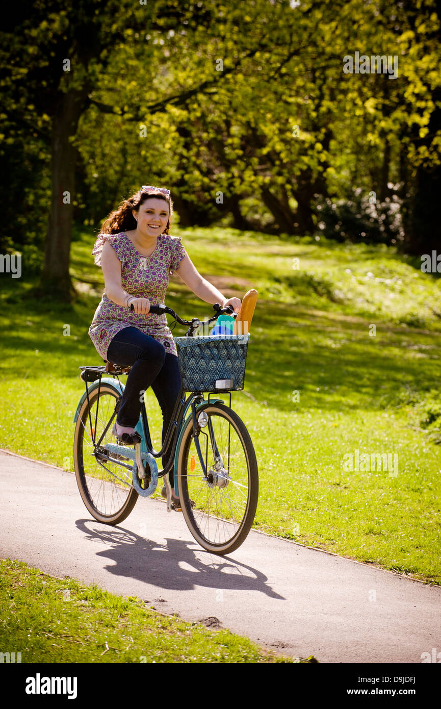 Junge kaukasische Frau, die im Sommer in einem britischen Park auf dem Fahrrad mit einer Flasche und einem Baguette im Korb unterwegs ist Stockfoto