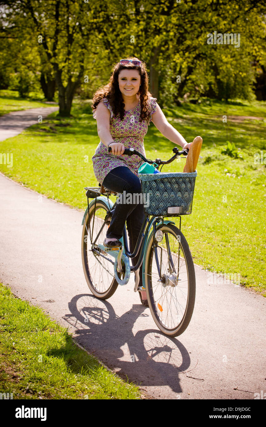 Junge kaukasische Frau, die im Sommer in einem britischen Park auf dem Fahrrad mit einer Flasche und einem Baguette im Korb unterwegs ist Stockfoto