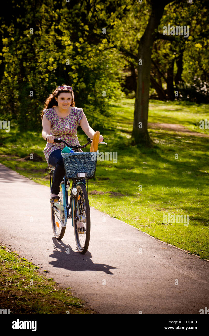 Junge kaukasische Frau, die im Sommer in einem britischen Park auf dem Fahrrad mit einer Flasche und einem Baguette im Korb unterwegs ist Stockfoto